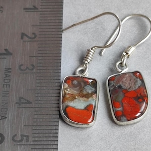 Puede incluir: Pendientes de plata con colgantes rectangulares de piedra colorida. Las piedras presentan tonos rojos, marrones y azules. Los pendientes miden aproximadamente 2,5 cm de largo, gancho incluido.