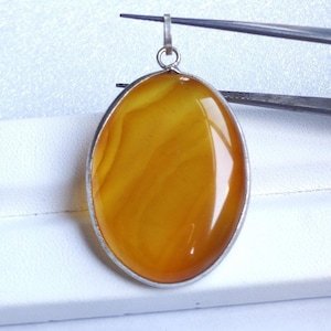 Op de afbeelding: Ovale barnsteen hanger met een zilverkleurig metalen frame. De hanger heeft een warme, goudgele kleur met natuurlijke patronen. Een kleine lus aan de bovenkant maakt het mogelijk om een ketting te bevestigen. Sieraden.