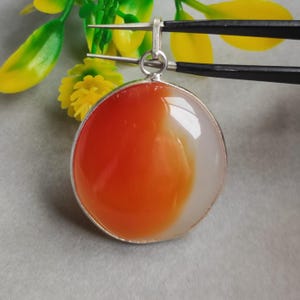 May include: A round pendant with a silver-colored bezel. The pendant features a polished stone with a gradient of orange to white. The pendant is being held by tweezers, with yellow and green foliage in the background.