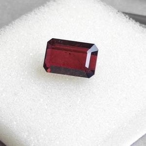 May include: A rectangular, faceted gemstone with a deep red hue. The gem is set against a white, textured background. The cut of the stone showcases its clarity and brilliance, highlighting its rich color.