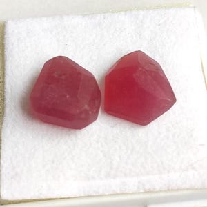 Könnte beinhalten: Zwei facettierte, rötlich-rosa Edelsteine mit unregelmäßigen Formen. Die Edelsteine werden auf einer weißen Oberfläche präsentiert, die ihre polierten Oberflächen und Farbvariationen zeigt. Diese Steine könnten für die Schmuckherstellung verwendet werden.