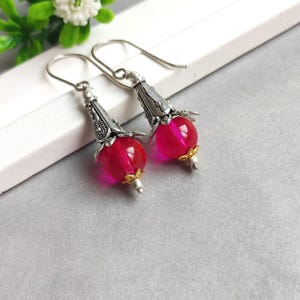 Puede incluir: Un par de pendientes colgantes con cuentas esféricas de color rosa intenso translúcido suspendidas de tapas de metal plateado ornamentadas. Cada pendiente incluye un pequeño detalle dorado en la base y un gancho plateado sencillo.