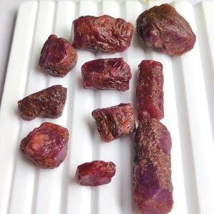 Könnte beinhalten: Eine Sammlung roher, ungeschliffener Rubin-Edelsteine in verschiedenen Formen und Größen. Die Edelsteine weisen einen tiefroten Farbton auf, wobei einige einen violetten Schimmer aufweisen. Die Steine sind auf einer weißen, geriffelten Oberfläche angeordnet, die ihre natürlichen Texturen und kristallinen Strukturen hervorhebt.