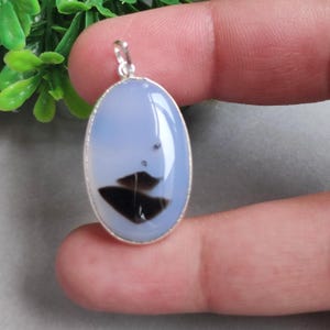 May include: An oval-shaped pendant featuring a smooth, translucent light blue stone with distinct dark brown, triangular inclusions. The gemstone is set in a simple silver-toned metal bezel frame with a small circular bail at the top for hanging.