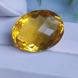 Könnte beinhalten: Ein facettierter, ovaler Edelstein mit einem warmen gelben Farbton. Die Oberfläche des Edelsteins reflektiert das Licht und erzeugt ein brillantes Farbspiel. Der Stein ist vor einem weißen Hintergrund platziert, was seine Klarheit und Brillanz hervorhebt.