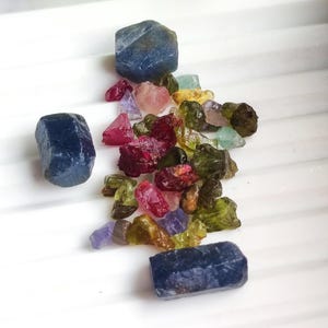 May include: A collection of raw gemstones in various colors, including deep blue, red, pink, green, and purple. The stones are scattered on a white surface, showcasing their natural textures and shapes. Ideal for jewelry making or collecting.