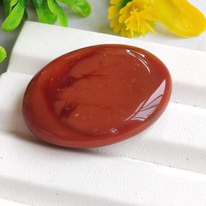 May include: A polished, oval-shaped, reddish-brown gemstone. The stone has a glossy surface and appears to be set on a white surface, with green leaves and yellow flowers in the background. The gemstone is approximately 2 inches in length.