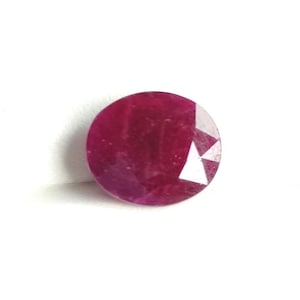 Könnte beinhalten: Ein facettierter, oval geformter Rubin-Edelstein. Der Edelstein hat eine tiefe, satte rote Farbe mit polierter Oberfläche und einigen Facetten auf der oberen rechten Seite. Der Rubin ist vor einem weißen Hintergrund platziert.