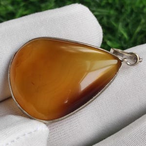 May include: A teardrop-shaped pendant with a polished, amber-colored stone set in a silver-toned metal frame. The stone has a gradient of colors, transitioning from light to dark brown. The pendant has a small loop for a chain.