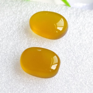 May include: Two oval-shaped, polished gemstones with a warm, golden-yellow hue. The stones have a smooth, glossy surface, reflecting light. The gemstones are set against a white, textured background.