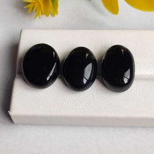 Könnte beinhalten: Drei polierte, oval geformte, schwarze Edelsteine werden auf einer weißen Oberfläche präsentiert. Die Steine haben eine glänzende Oberfläche, die das Licht reflektiert. Die Edelsteine werden wahrscheinlich für Schmuck oder Dekorationszwecke verwendet.