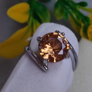 Könnte beinhalten: Ein silberner Ring mit einem großen, runden, facettierten, hellorangen Edelstein. Der Edelstein wird von vier Krappen gehalten. Der Ring hat ein einzigartiges, geschwungenes Design.