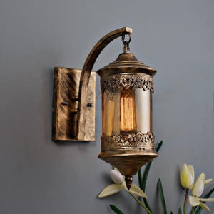 Könnte beinhalten: Eine Wandleuchte im antiken Stil mit goldener Oberfläche. Die Leuchte hat ein dekoratives, verziertes Design mit einem transparenten Glaszylinder und einer warm leuchtenden Glühbirne. Die Leuchte ist auf einer rechteckigen Basis montiert und hat einen gebogenen Arm.