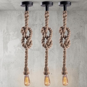 Pode incluir: Três luminárias pendentes com tema náutico. Cada luminária apresenta um design de corda marrom com um nó decorativo, uma montagem de teto preta e uma lâmpada Edison de estilo vintage. As luzes pendem contra uma parede de concreto cinza.