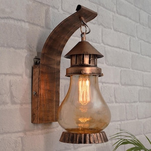Peut inclure: Applique murale de style vintage avec un abat-jour en verre ambré chaud et un cadre en métal décoratif. Le luminaire est équipé d'une ampoule de style Edison et est monté sur un support en bois. Le design général est rustique et industriel.