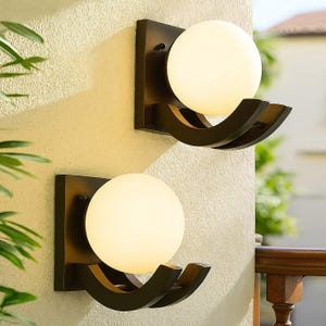 May include: Two outdoor wall sconces with a dark brown square backplate and a curved, decorative arm holding a spherical, frosted glass globe. The sconces are mounted on a light beige stucco wall.