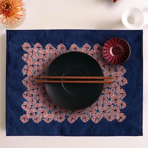 Op de afbeelding: Een gedekte tafel met een marineblauwe placemat met een koraalrood bloemmotief. Een zwart bord met houten eetstokjes ligt op de placemat. Een klein rood schaaltje en een bloemstuk zijn ook zichtbaar.