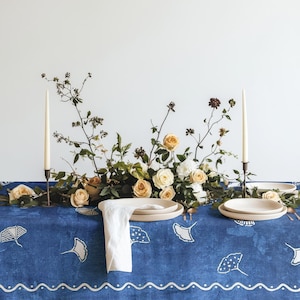 Peut inclure: Une table dressée avec une nappe bleue ornée d'un motif de feuilles blanches. La table est décorée d'une composition florale de roses et de verdure, de deux grandes bougies et d'assiettes beiges. Une serviette blanche est pliée sur la table.