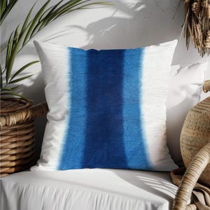 Peut inclure: Coussin carré blanc avec une large bande verticale bleu indigo. La bande bleue présente un dégradé de couleurs. Le coussin est probablement fait d'un tissu texturé, pour la décoration.