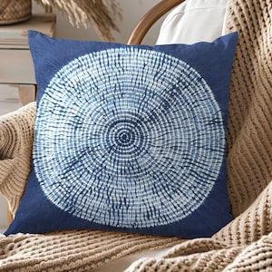 Könnte beinhalten: Ein quadratisches, marineblaues Zierkissen mit einem weißen, kreisförmigen Muster, das an einen Baumring erinnert. Das Kissen liegt auf einem Holzstuhl mit einem beigefarbenen Stricküberwurf.