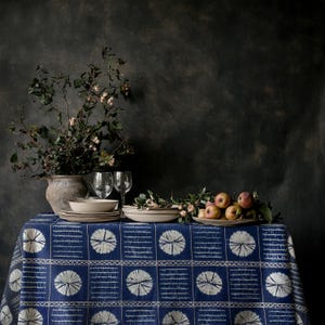 May include: A table set for dining, with a blue and white patterned tablecloth. There are plates, glasses, and a vase with foliage. A plate of apples is also present. The backdrop is a dark, textured wall.