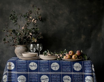 May include: A dining table set with a blue and white patterned tablecloth, featuring plates, glasses, and a vase with greenery. Apples are arranged on a plate. The background is a dark, textured wall.