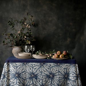 Pode incluir: Uma mesa de jantar com uma toalha de mesa estampada em azul e branco. Pratos bege, taças de vinho e um vaso com vegetação estão dispostos sobre a mesa. Um prato de maçãs adiciona um toque de cor à cena.