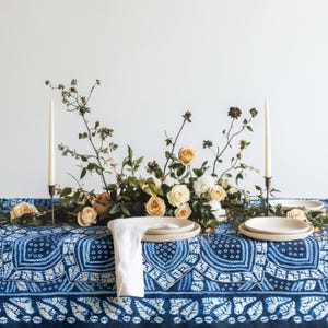 Peut inclure: Une table élégante avec une nappe à motifs bleus et blancs. Des assiettes crème, une serviette blanche et un arrangement floral de roses et de verdure ornent la table. De hautes bougies dans des bougeoirs créent une atmosphère sophistiquée.
