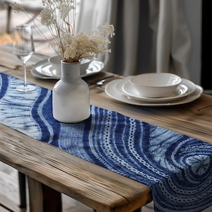 May include: A wooden table is set with a blue and white patterned table runner. A white vase holds dried flowers. White plates, a bowl, and a wine glass are also on the table, creating a serene dining scene.