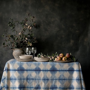 Puede incluir: Una mesa con un mantel estampado en azul y blanco. Un jarrón con vegetación y dos copas de vino están sobre la mesa. Hay platos y un cuenco apilados, y también hay un plato con manzanas.