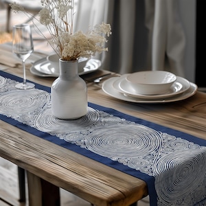 May include: A navy blue table runner with a white spiral design, set on a wooden table. A white vase with dried flowers, white plates, and a wine glass complete the dining table setting.