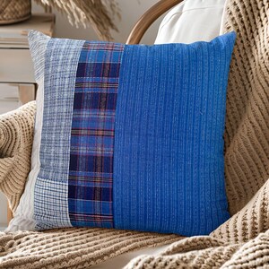 May include: Square decorative pillow with a patchwork design. The pillow features sections of blue fabric, including a plaid pattern and a solid textured section. The pillow is placed on a wooden chair with a beige knitted throw.