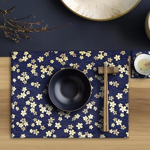 Può includere: Un sottopiatto blu navy con un motivo floreale dorato, una ciotola abbinata e bacchette. Il sottopiatto presenta un motivo a onde. Sono presenti anche una piccola ciotola bianca con tè e un poggia bacchette. L'allestimento è su una superficie di legno.