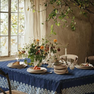 May include: A dining table set for a meal, featuring a navy blue tablecloth with a white patterned border. The table is set with plates, bowls, glasses, silverware, and a floral centerpiece with orange and yellow flowers. Two lit candles add to the ambiance.