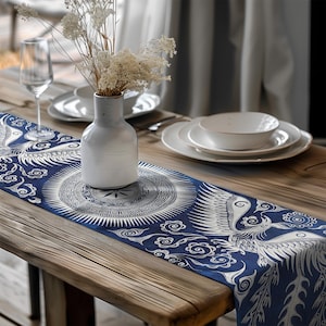 Peut inclure: Chemin de table bleu et blanc avec un motif de phénix. Le chemin de table est posé sur une table en bois, avec un vase blanc et des fleurs séchées au centre. Des assiettes blanches et un verre à vin sont également visibles.