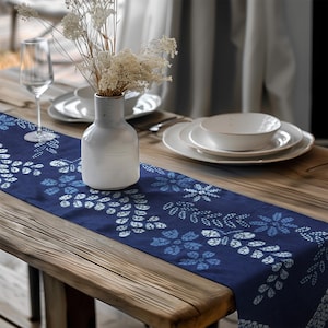 May include: A navy blue table runner with a white and light blue floral pattern. The runner is placed on a wooden table, alongside white plates, a vase, and a wine glass. The vase holds dried flowers.
