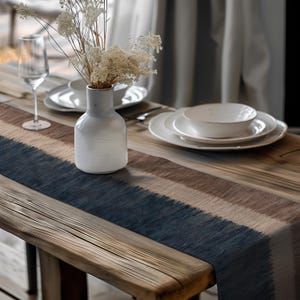 May include: A wooden table set with a blue, brown, and beige striped table runner. A white vase holds dried flowers. White plates, a bowl, and a wine glass are also on the table, creating a neutral and natural aesthetic.