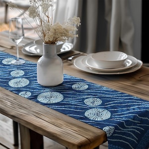 Puede incluir: Camino de mesa azul con un diseño circular blanco y líneas onduladas. Un jarrón blanco con flores secas está sobre el camino de mesa, junto con platos blancos y una copa de vino. Sobre una mesa de madera.
