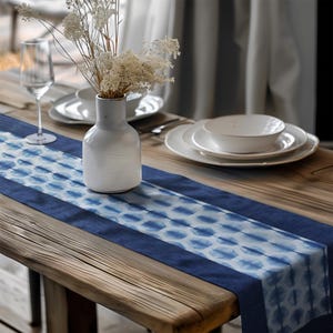 Könnte beinhalten: Ein rustikaler Holztisch ist mit einem blau-weiß gemusterten Tischläufer gedeckt. Eine weiße Vase enthält Trockenblumen, und weiße Teller und ein Weinglas sind auf dem Tisch angeordnet. Der Läufer hat ein Batik-Design.