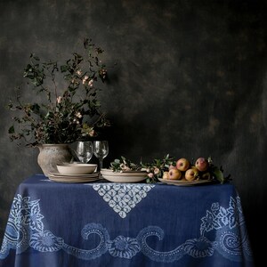 Puede incluir: Naturaleza muerta con un mantel azul con bordes estampados en blanco, con platos, vasos y un jarrón con vegetación. Las manzanas están dispuestas en un plato. El fondo es una pared oscura y texturizada.