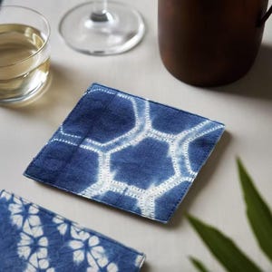Könnte beinhalten: Quadratischer blau-weißer Stoffuntersetzer mit Wabenmuster. Der Untersetzer besteht aus strukturiertem Stoff und liegt auf einer hellen Oberfläche. Ein Glas Flüssigkeit und eine dunkelbraune Tasse befinden sich im Hintergrund.