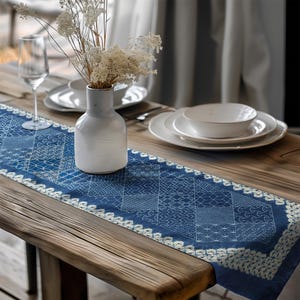 Può includere: Un runner da tavolo blu con bordatura in pizzo bianco e motivi geometrici è appoggiato su un tavolo di legno. Un vaso bianco con fiori secchi, piatti bianchi e un calice sono anche sul tavolo.