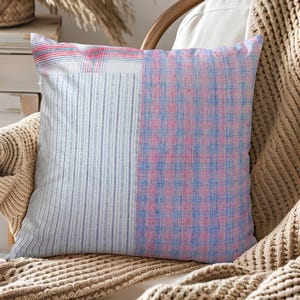 Peut inclure: Coussin carré décoratif avec un motif patchwork. Un côté présente des rayures verticales bleues et blanches, l'autre un motif à carreaux roses et bleus. Le coussin est posé sur une chaise en bois avec un plaid beige tricoté.