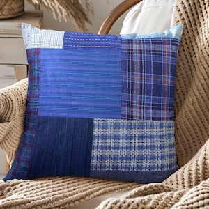 Puede incluir: Cojín decorativo cuadrado con un diseño de patchwork en tonos azules. El cojín presenta varios patrones de tela, incluyendo rayas y cuadros. Descansa sobre una silla de madera con una manta de punto beige.