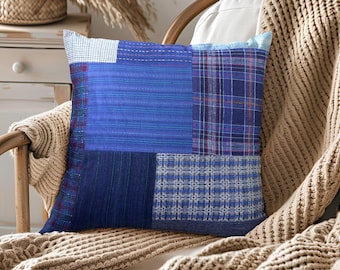 Könnte beinhalten: Quadratisches Zierkissen mit Patchwork-Design in Blautönen. Das Kissen weist verschiedene Stoffmuster auf, darunter Streifen und Karos. Es liegt auf einem Holzstuhl mit einem beigefarbenen Strickwurf.
