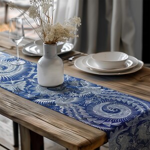 May include: A blue and white patterned table runner with a floral design is laid on a wooden table. A white vase with dried flowers sits on the runner, with white plates and a wine glass in the background.