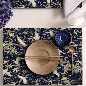 Puede incluir: Una mesa puesta con un mantel individual azul marino con un patrón de grullas blancas y olas doradas. Un plato grande de madera, palillos chinos y un cuenco azul pequeño están sobre el mantel. También se ven una cuchara de madera y una flor morada.