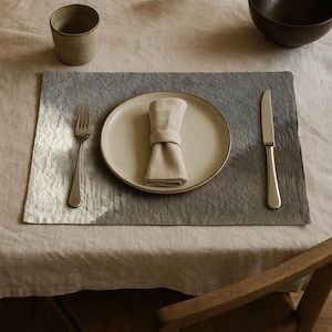 Puede incluir: Un cubierto con un plato beige, una servilleta doblada, un tenedor y un cuchillo sobre un mantel individual gris y blanco. También se ven una taza de cerámica pequeña y un cuenco oscuro. La mesa está cubierta con un mantel beige.