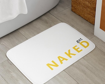 Op de afbeelding: Witte rechthoekige badmat met afgeronde hoeken. De mat heeft de woorden "get NAKED" in het geel gedrukt. De mat ligt op een lichte houten vloer naast een bad en een rieten mand.