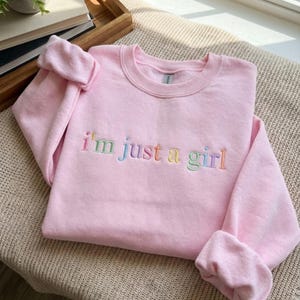Może przedstawiać: Jasnoróżowa bluza z haftowanym napisem "i'm just a girl" w tęczowych kolorach. Bluza z okrągłym dekoltem ma długie rękawy, z podwiniętymi mankietami. Ubranie jest starannie złożone na neutralnej powierzchni.